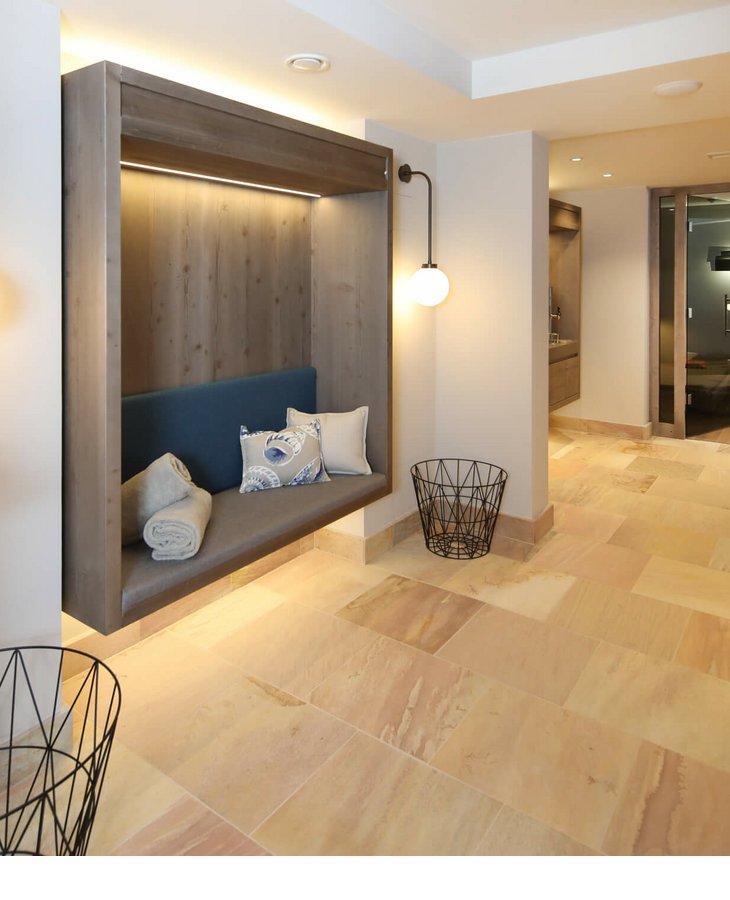 Moderner Hotelflur mit Holzbank, Kissen, Decken, warmem Licht und Blick in einen gemütlichen Lounge-Bereich.