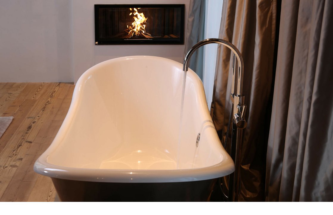 Freistehende Badewanne mit fließendem Wasser, Holzfußboden, Kamin im Hintergrund, elegante Vorhänge.