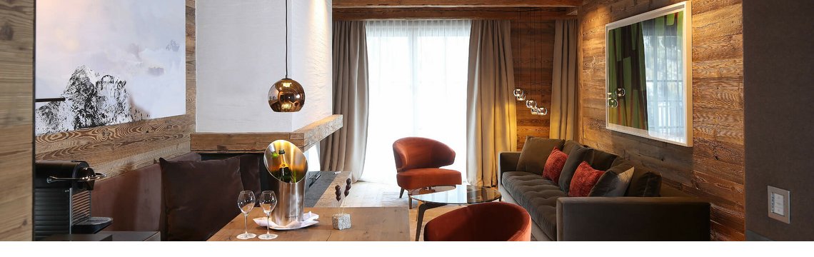 Gemütliches Hotelzimmer mit Holzwänden, eleganter Sitzecke, Esstisch, Kunstwerk und moderner Beleuchtung.