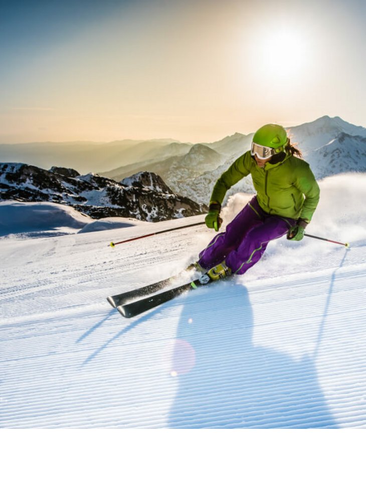 Skifahrer in grüner Jacke fährt dynamisch eine schneebedeckte Piste hinab, umgeben von Berglandschaft und Sonnenschein.
