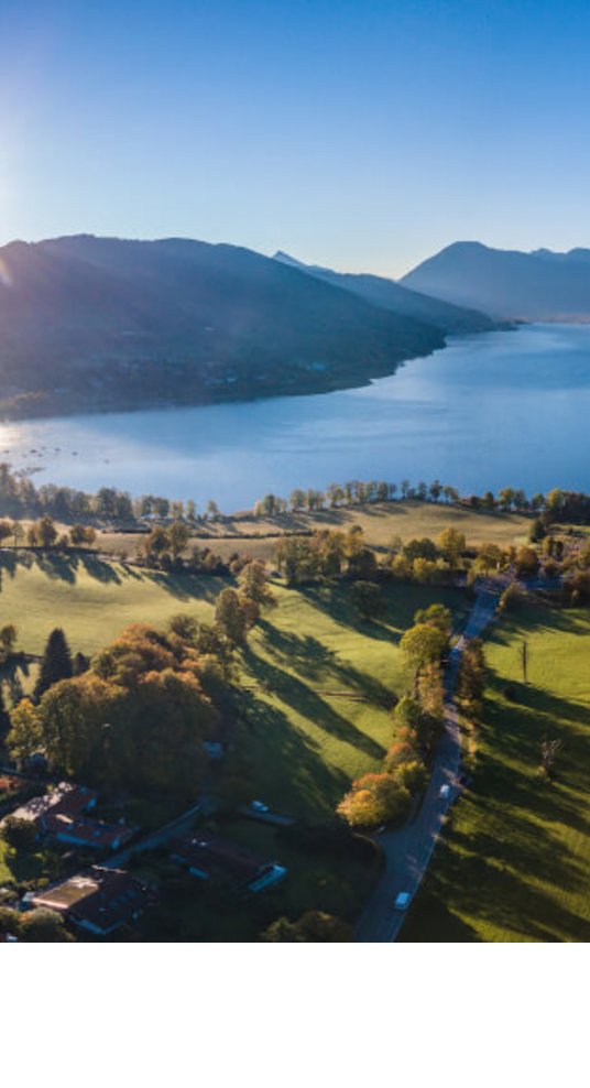 Luftaufnahme eines Sees mit umliegenden grünen Wiesen und Bergen im Hintergrund bei Sonnenaufgang.