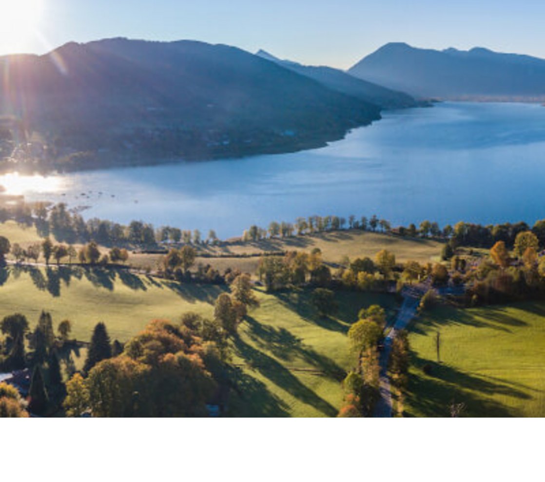 Severin*s Resort & Spa Tegernsee Luftaufnahme eines Sees mit umliegenden grünen Wiesen und Bergen im Hintergrund bei Sonnenaufgang.
