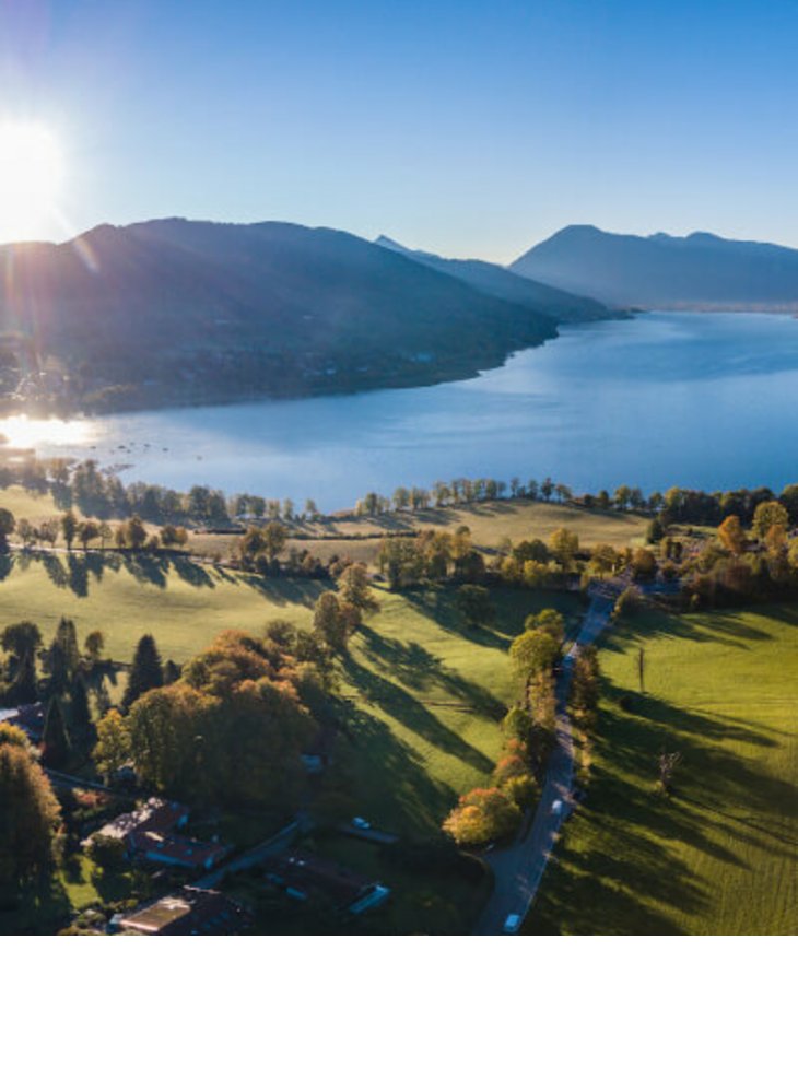 Luftaufnahme eines Sees mit umliegenden grünen Wiesen und Bergen im Hintergrund bei Sonnenaufgang.