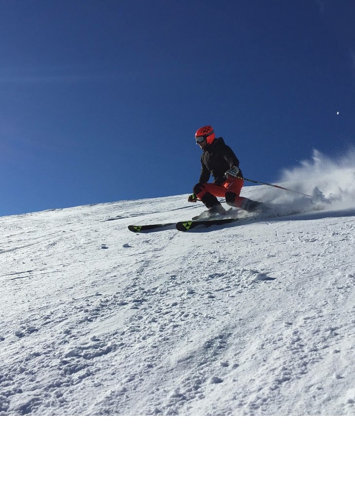 Skifahrer in roter Ausrüstung fährt dynamisch eine schneebedeckte Piste hinab, blauer Himmel im Hintergrund.