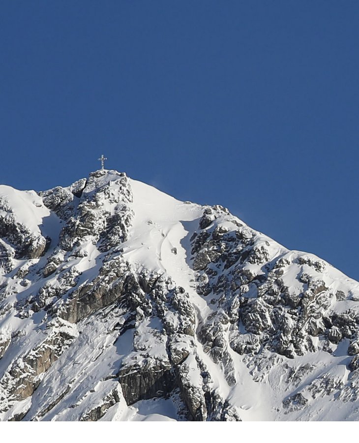Schneebedeckter Berggipfel mit Gipfelkreuz vor klarem, blauem Himmel.