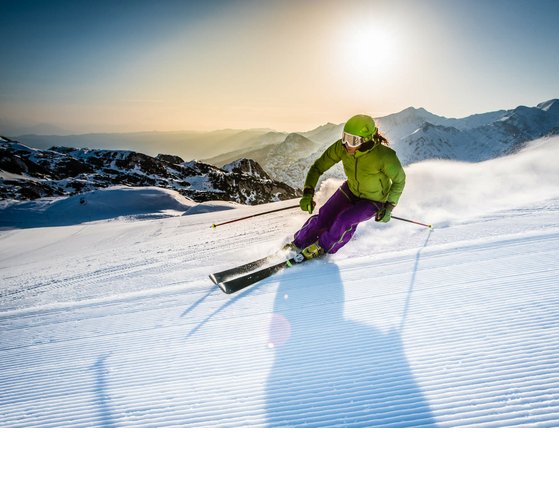 Skifahrer in grüner Jacke fährt bei Sonnenschein eine verschneite Piste hinab, Berge im Hintergrund.