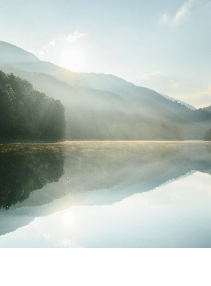 Bergsee im Morgenlicht, umgeben von bewaldeten Hügeln, mit ruhiger Wasseroberfläche und sanftem Nebel.