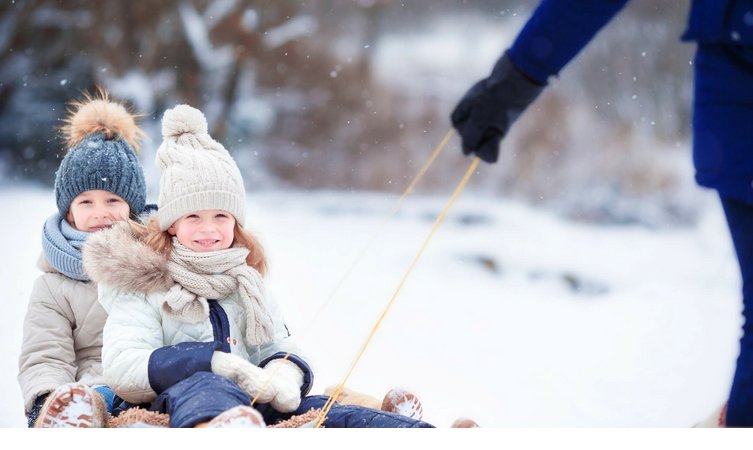 Zwei Kinder in Winterkleidung lächeln auf einem Schlitten im Schnee, während eine Person sie zieht.