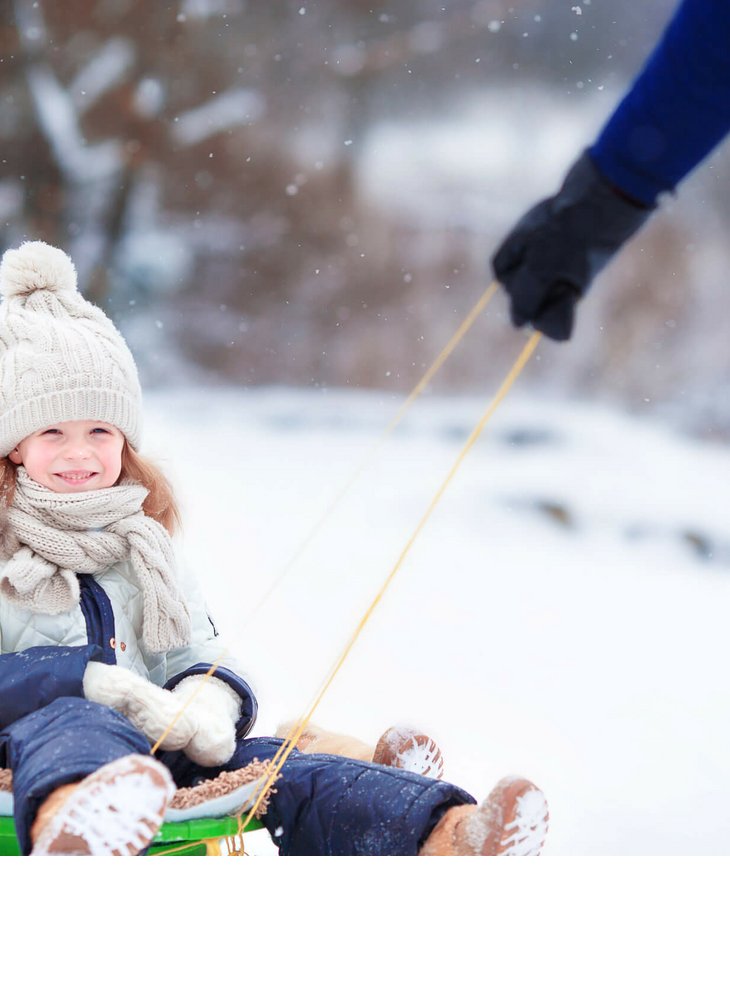 Zwei Kinder in Winterkleidung lächeln auf einem Schlitten im Schnee, während eine Person sie zieht.