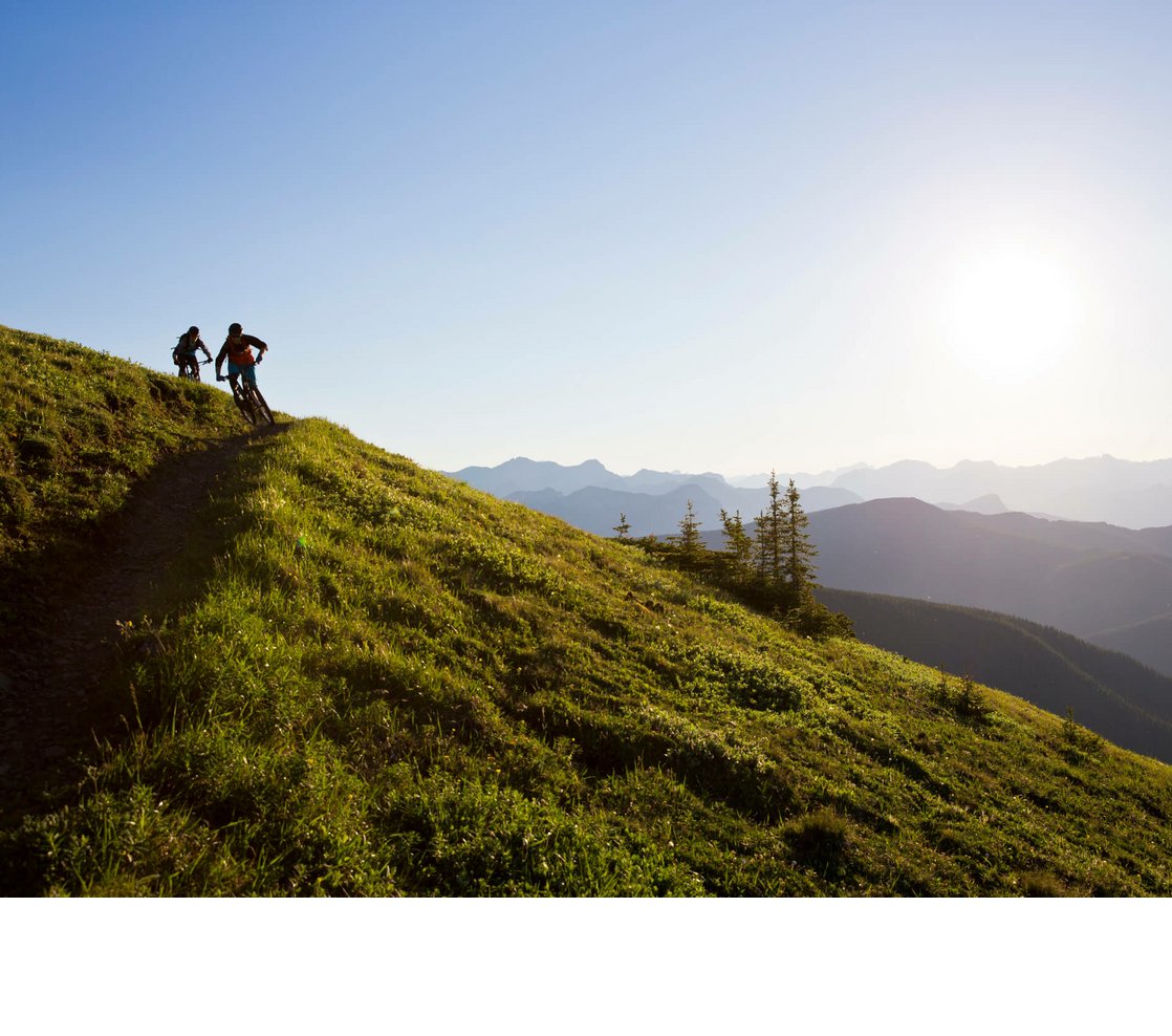 Radfahren in den Bergen | Severin*s The Alpine Retreat Zwei Mountainbiker fahren bei Sonnenuntergang auf einem grünen Berghang mit Alpenpanorama.
