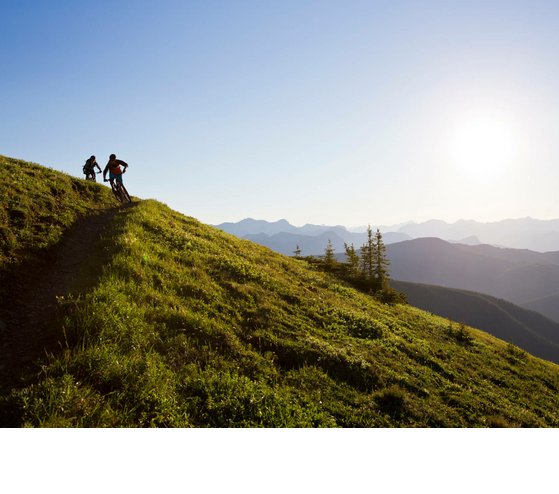 Radfahren in den Bergen | Severin*s The Alpine Retreat Zwei Mountainbiker fahren bei Sonnenuntergang auf einem grünen Berghang mit Alpenpanorama.