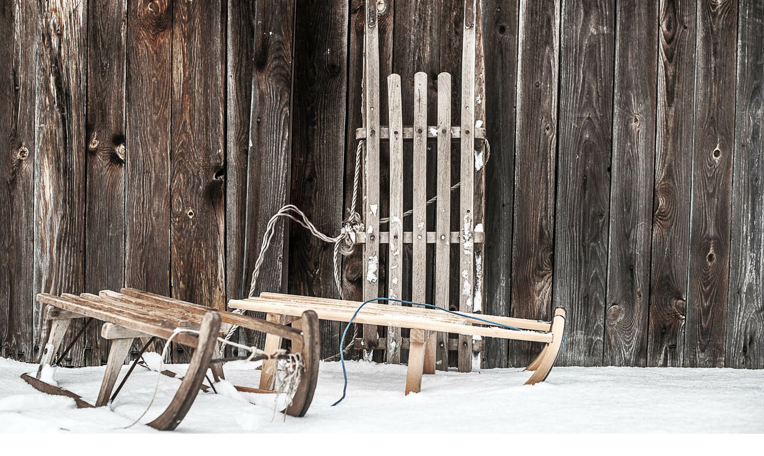 Zwei hölzerne Schlitten lehnen an einer rustikalen Holzwand im Schnee.