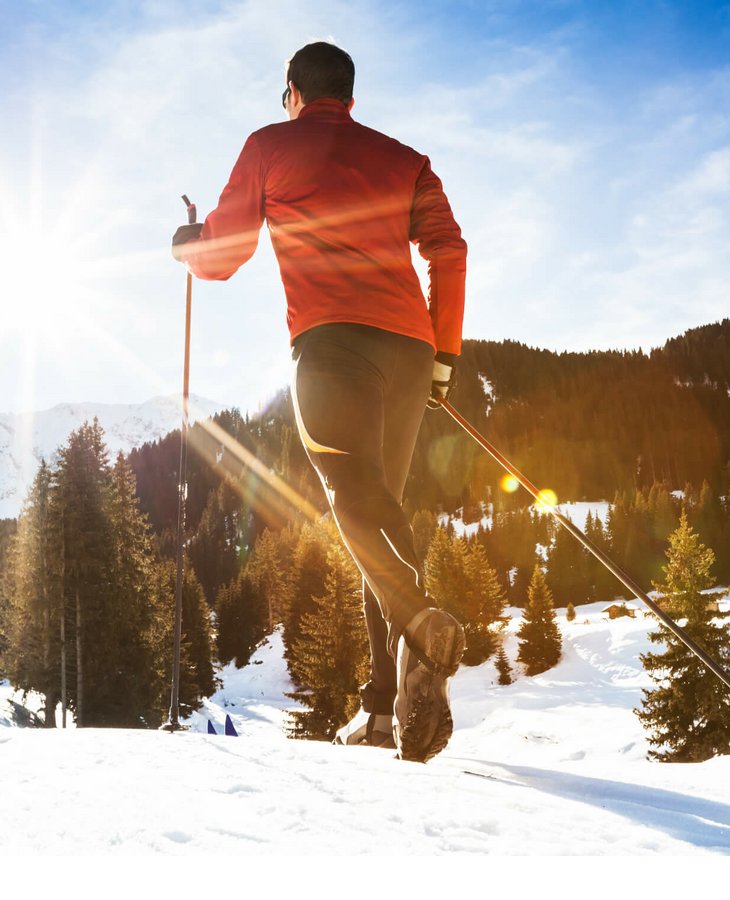 Person beim Langlaufen im Schnee, mit Sonne im Hintergrund und verschneiten Bergen und Wäldern.