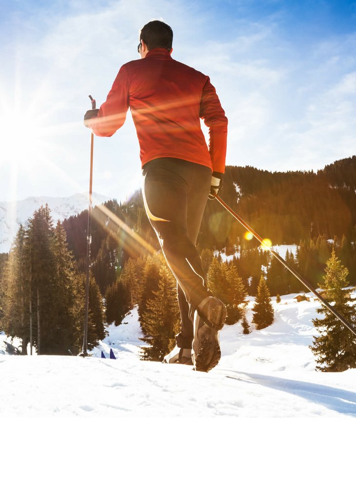 Nordic Walking im Winter | Severin*s The Alpine Retreat Person beim Langlaufen im Schnee, mit Sonne im Hintergrund und verschneiten Bergen und Wäldern.