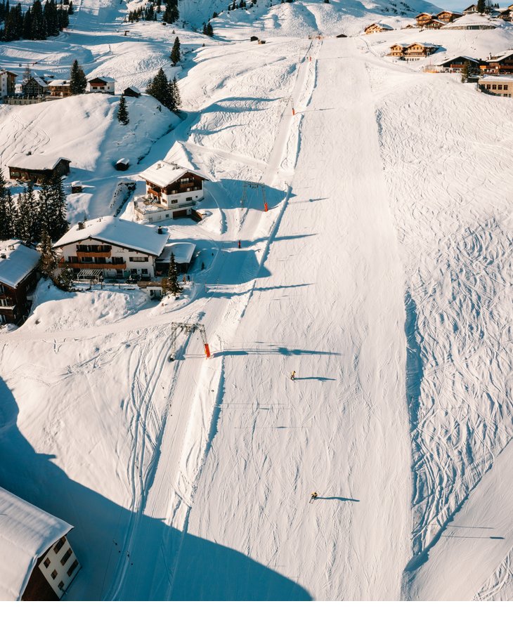Verschneite Skipiste mit Skifahrern, umgeben von Chalets und Bergen unter blauem Himmel.
